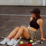 Young woman sitting on a tennis court with rackets and tennis balls, enjoying a relaxed moment outdoors.